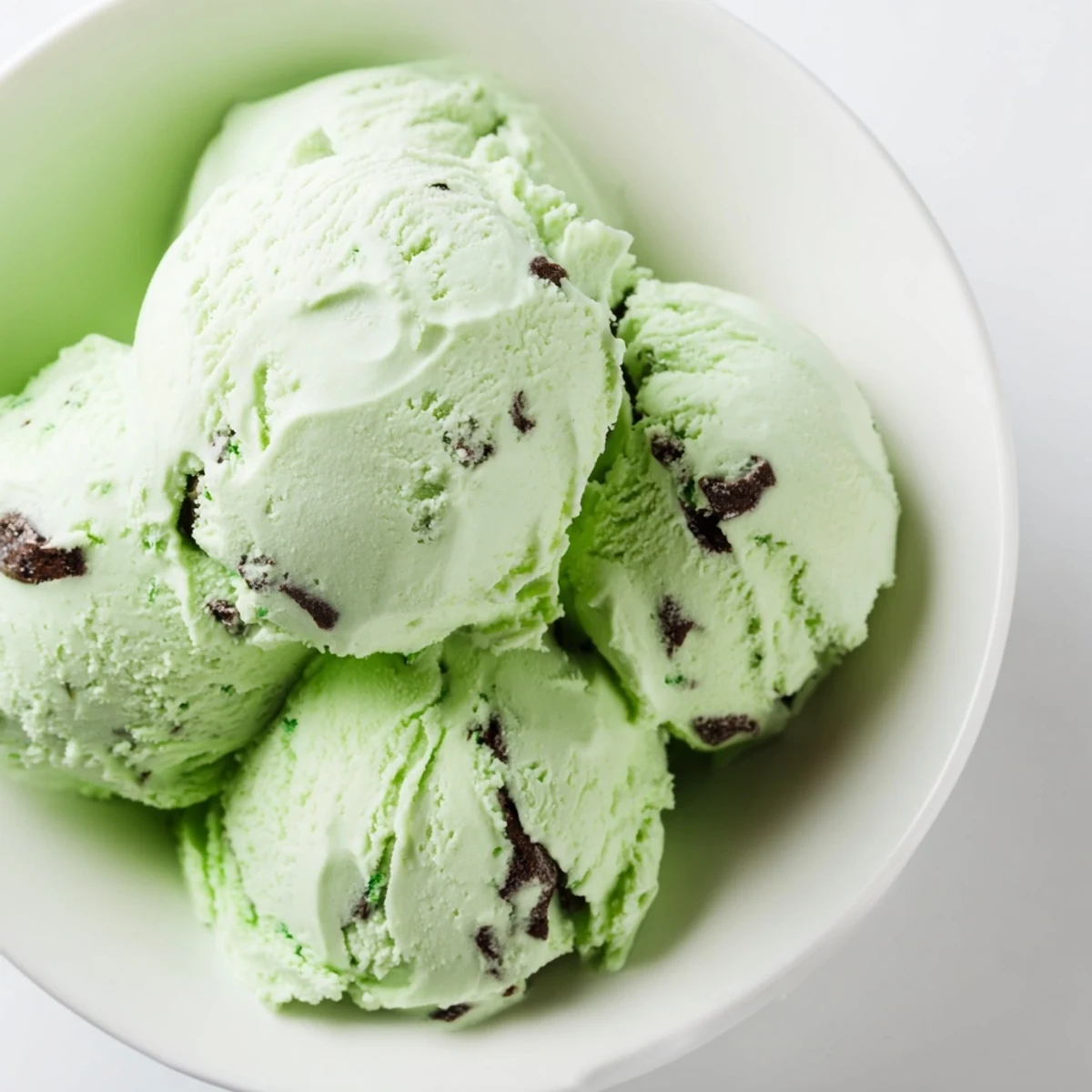 Scoops of Green Mint Chip Ice Cream melting slightly on a dark plate, sprinkled with extra chocolate chips and mint sprigs for an inviting look.