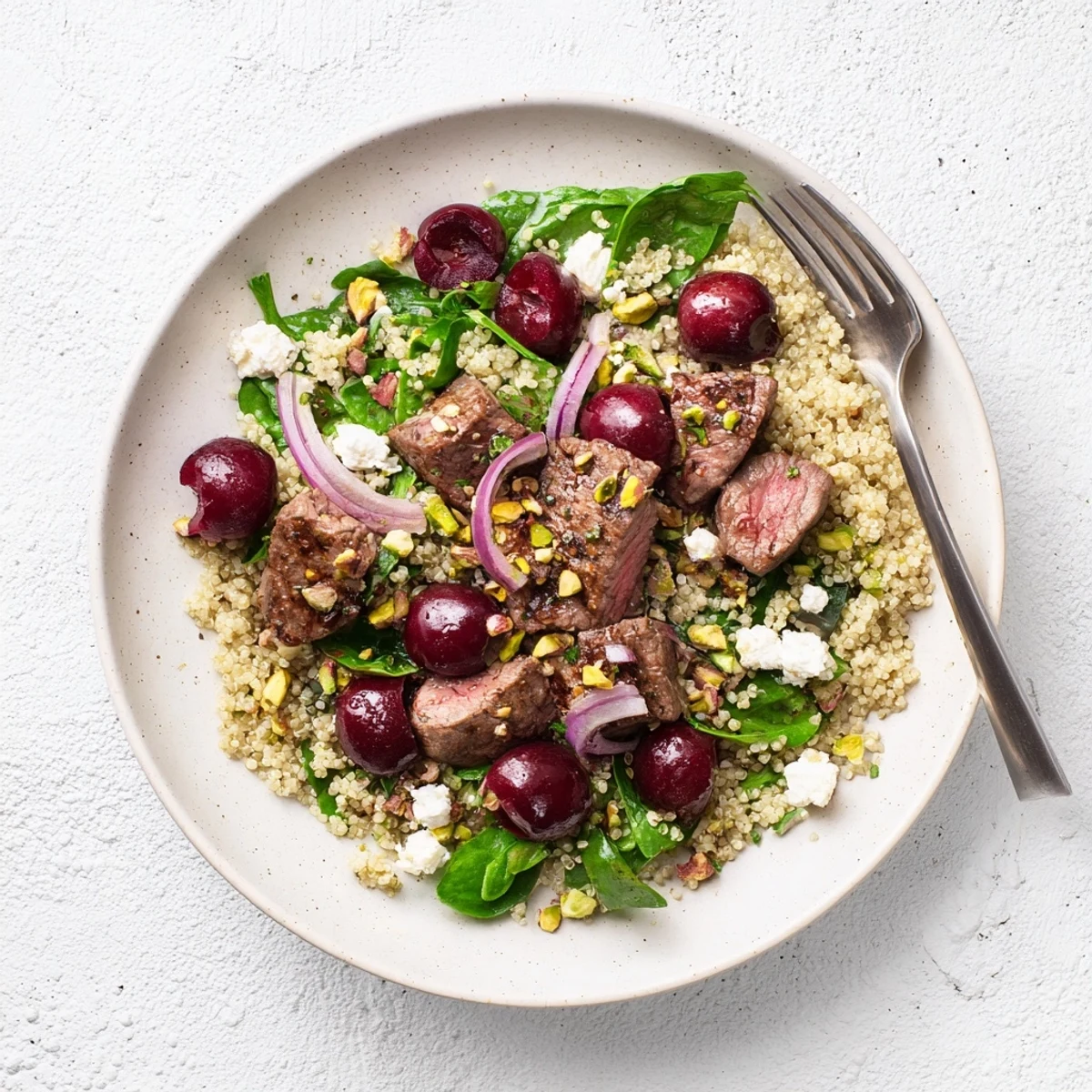 Colorful cherry quinoa salad with lamb topped with feta, fresh herbs, and toasted pistachios on a white plate