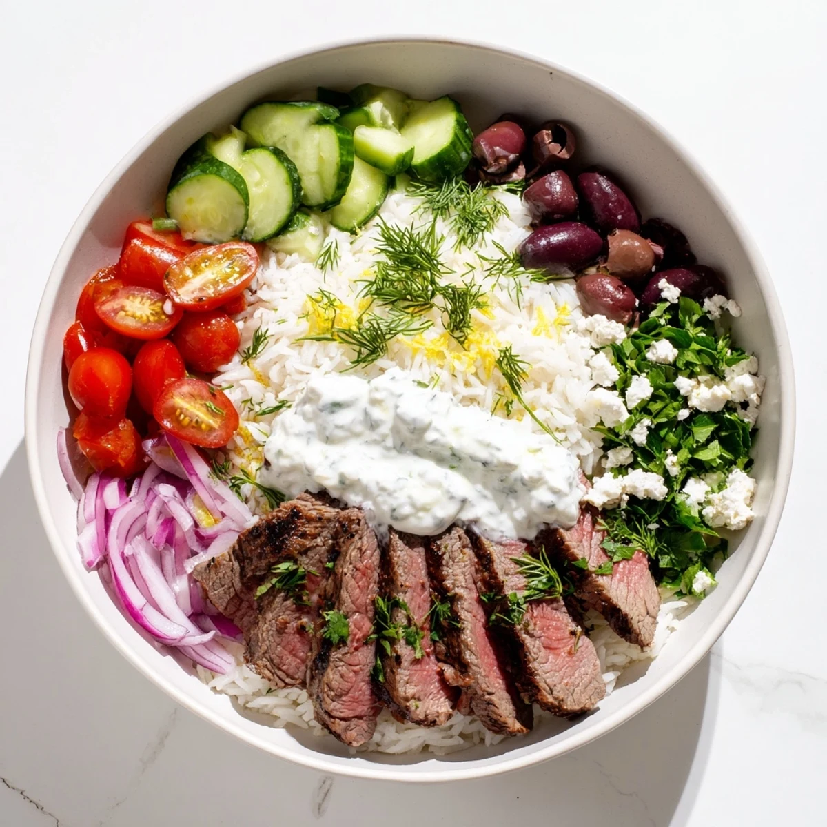Sliced steak over seasoned rice with tomatoes, cucumber, olives, and drizzled yogurt sauce in Mediterranean steak bowl