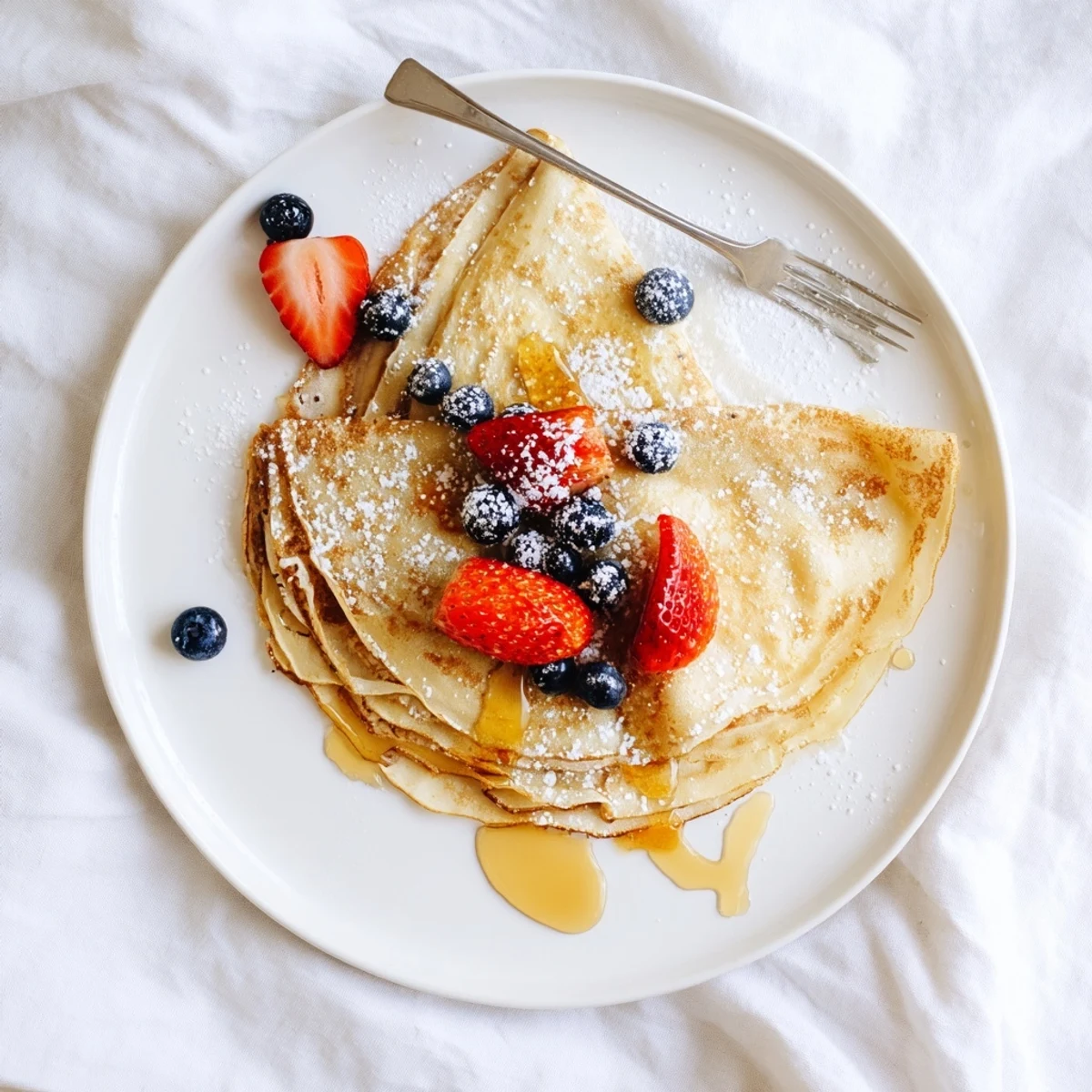 Stack of thin French crepes with honey drizzle garnished with fresh berries and powdered sugar