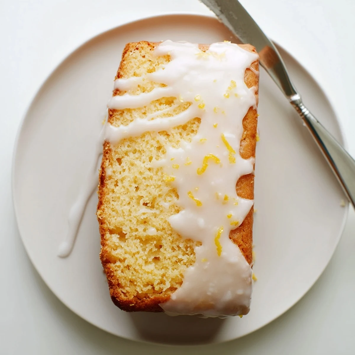Moist lemon loaf cake slice topped with tangy glaze beside fresh lemons on white plate