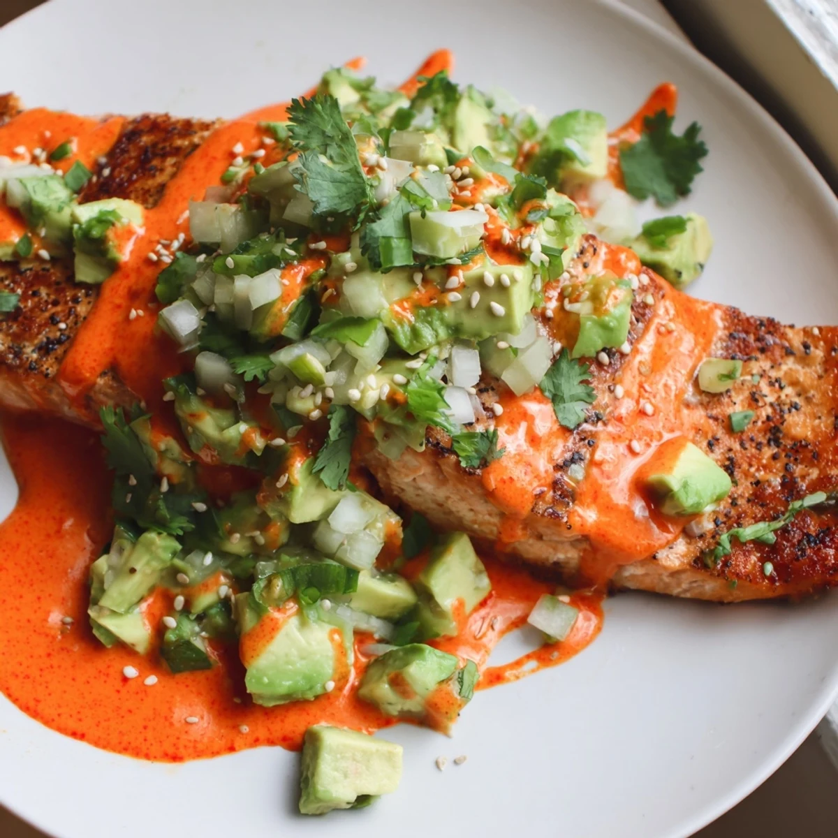 Crispy baked Bang Bang Salmon drizzled with spicy cream sauce and fresh avocado cucumber salsa on a white plate